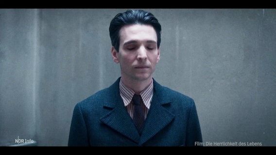 Ein Mann mit dunklen, zurückgekämmten Haaren steht vor einer grauen Wand. Sein Blick ist gesenkt, seine Augen sind geschlossen. © Screenshot 