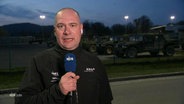 Ein Mann steht auf militärischem Gelände und spricht in ein Mikrofon. © Screenshot 