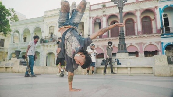 Ein junger Mann mit vielen Tattoos macht Brakedance auf einem öffentlichen Platz. © Screenshot 