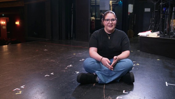 Eine Schülerin im Teenager-Alter sitzt im Schneidersitz auf dem schwarzen Boden einer Bühne und gibt ein Interview. © Screenshot 