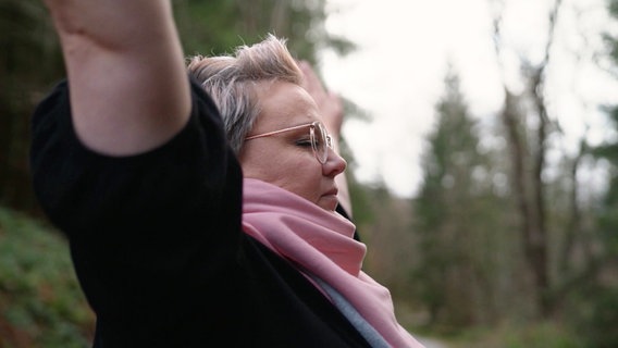 Eine blonde Frau mit Brille streckt mit geschlossenen Augen die arme in die Luft © Screenshot 