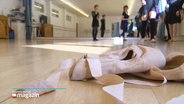 Ballettschuhe auf dem Parkett eines Saals mit mehreren Personen im Hintergrund. © Screenshot 