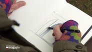 Eine Person malt eine Situationsaufnahme mit Bleistift in ein Notizbuch. © Screenshot 