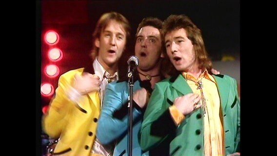 Ein Männer-Trio steht in bunten Anzügen auf der Bühne und singt ins Mikrofon. © Screenshot 