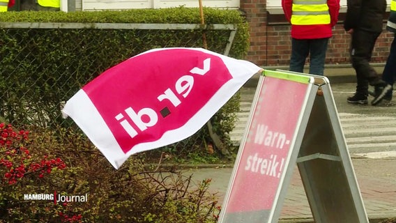 Ein Dreiecksständer kündigt den Warnstreik an. Daran befestigt flattert eine Ver.di-Fahne im Wind. © Screenshot 