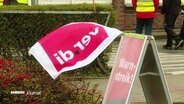 Ein Dreiecksständer kündigt den Warnstreik an. Daran befestigt flattert eine Ver.di-Fahne im Wind. © Screenshot 