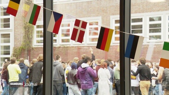 Eine Girlande mit den Flaggen verschiedener europäischer Länder hängt vor einer Fensterscheibe. Draußen steht eine große Gruppe junger Menschen. © Screenshot 