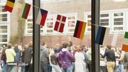 Eine Girlande mit den Flaggen verschiedener europäischer Länder hängt vor einer Fensterscheibe. Draußen steht eine große Gruppe junger Menschen. © Screenshot 