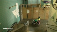 Ein Rollstuhlfahrer vor der vernagelten Baustelle eines Fahrstuhls an einer S-Bahn-Station. Im Vordergrund ist eine Treppe und Rolltreppe im Bild. © Screenshot 
