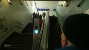 Die Kamera filmt über die Schulter einer Person am Fuße einer Treppe und einer Rolltreppe an einer S-Bahn-Station. Die Person schaut nach oben in Richtung Gleis. © Screenshot 