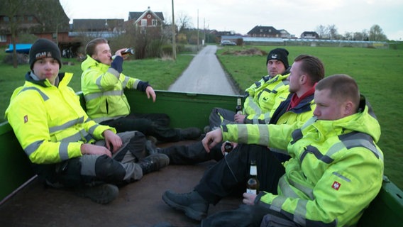 Fünf junge Männer in neongelben Jacken sitzen auf der Ladefläche eines Fahrtzeugs und trinken Bier. © Screenshot 