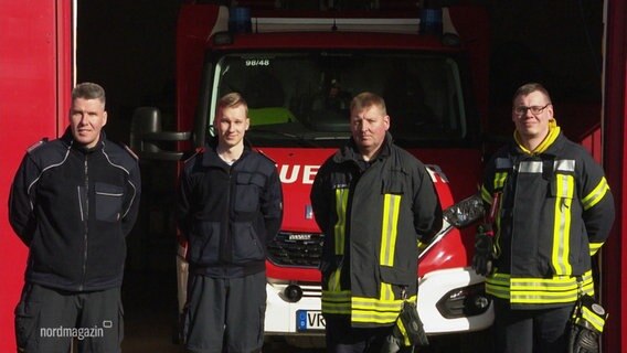 Vier Feuerwehrleute in Uniform vor der offenen Garage mit dem Feuerwehrfahrzeug. © Screenshot 