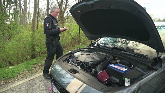 Ein Polizist steht an einem Auto mit offener Kühlerhaube. © Screenshot 