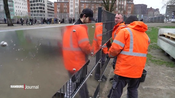 Mehrere Personen in oranger Arbeitskleidung bringen Sichtschutzblenden an einen Zaun vor der Drogenberatungsstelle "Drob Inn" an. © Screenshot 