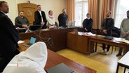 Szene im Gerichtssaal: Alle Beteiligten wie Richter und Verteidiger stehen, der Angeklagte sitzt, im Vordergrund im Anschnitt zu sehen. © Screenshot 