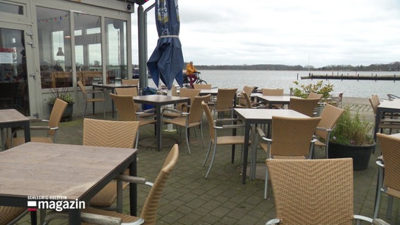 Unbesetzte Stühle und Tische draußen vor einem Café am Wasser. © Screenshot 