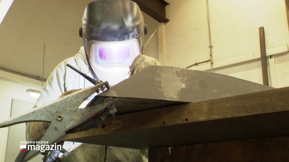 Eine Person mit Schutzkleidung und Schweißhelm arbeitet an einem Werkstück aus Metall in einer Werkstatt. © Screenshot 