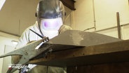 Eine Person mit Schutzkleidung und Schweißhelm arbeitet an einem Werkstück aus Metall in einer Werkstatt. © Screenshot 