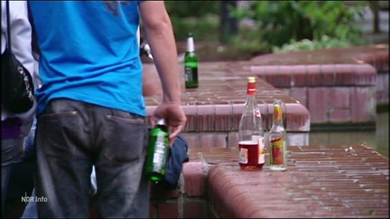 Eine Person mit einer Bierflasche in der Hand von hinten; daneben stehen auf einer Mauer Alkoholflaschen, teilweise mit Resten. © Screenshot 