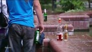 Eine Person mit einer Bierflasche in der Hand von hinten; daneben stehen auf einer Mauer Alkoholflaschen, teilweise mit Resten. © Screenshot 