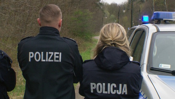 Ein Polizist und eine Polizisten von hinten fotografiert. Sie stehen neben einem Streifenwagen. © Screenshot 