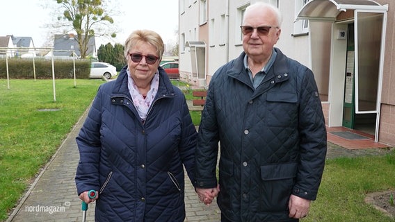 Ein älteres Ehepaar steht händchenhaltend vor der Kamera, im Hintergrund ist ein Mehrfamilienhaus zu sehen. Beide Personen tragen Jacken und Sonnenbrillen. © Screenshot 
