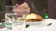 Gläser und eine Mahlzeit auf einem Tisch, daneben sitzt eine  Person. © Screenshot 