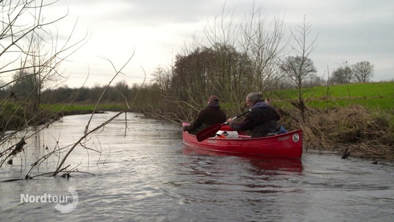 Zwei Männer sitzen in einem roten Kanu und paddeln über einen kleinen Fluss. © Screenshot 