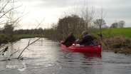 Zwei Männer sitzen in einem roten Kanu und paddeln über einen kleinen Fluss. © Screenshot 