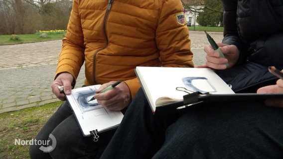 Zwei Menschen sitzen draußen auf kleinen Hockern und zeichnen in ihre Skizzenbücher. © Screenshot 