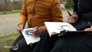 Zwei Menschen sitzen draußen auf kleinen Hockern und zeichnen in ihre Skizzenbücher. © Screenshot 