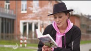 Annie Heger sitzt mit einem großen schwarzen Hut draußen und liest aus ihrem Buch. © Screenshot 