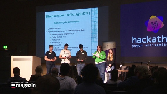 Die Bühne des Hackathon gegen Antisemitismus, auf der Menschen mit Mikrofonen stehen. © Screenshot 