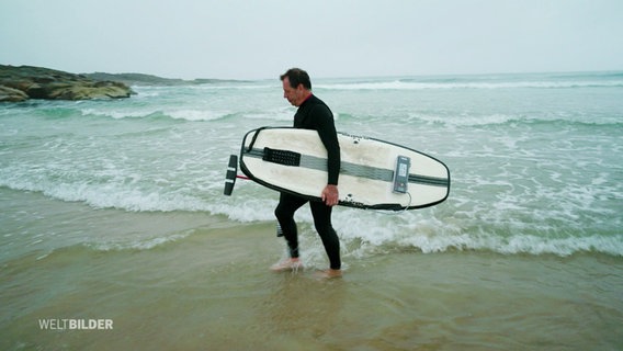 Ein Mann in dunklem Neoprenanzug läuft mit einem Surfbrett unter dem Arm aus dem Meer heraus. © Screenshot 