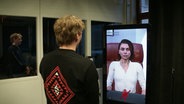 Ein Mann steht vor einem Bildschirm, auf dem eine compter-animierte Frau zu sehen ist. © Screenshot 