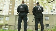 Aus der Froschperspektive: Zwei mit Schutzkleidung, Schlagstöcken und Waffen ausgerüstete Polizeibeamte stehen vor einem Plattenbau. © Screenshot 