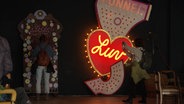 Menschen stehen neben einer Instalation mit Neonlichtern. In einem rotn Herz leuchtet der Schriftzug "Love". © Screenshot 