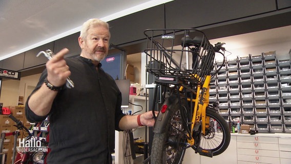 Ein Mann steht in einer Fahrradwerkstatt und hält Werkzeuge in der Hand. © Screenshot 