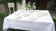 Ein Tisch ist mit weißer Tischdecke und geblümten Tassen und Tellern gedeckt. In der Mitte stehen Blumen. © Screenshot 
