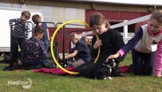 Einige Kinder sitzen mit Spielgeräten und Welpen auf einer Wiese im Hof. © Screenshot 