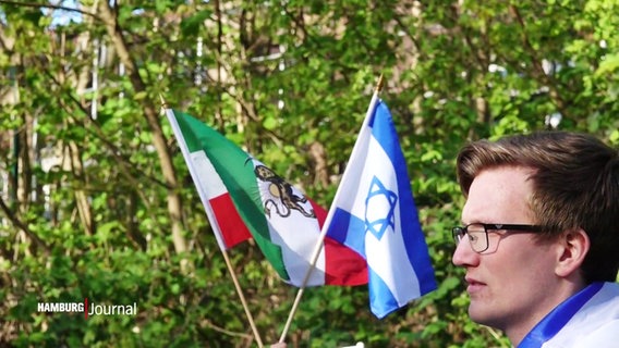 Die iranische und die israelische Flagge nebeneinander. Von rechts steht ein Mann im Profil. Er schaut zu etwas außerhalb des linken Bildrandes. © Screenshot 