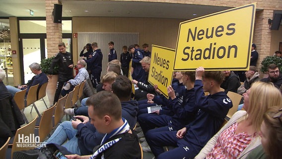 Jugendliche in blauen Trainingsanzügen fordern mit gelben Schildern ein "neues Stadion". © Screenshot 