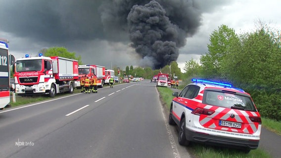 An einer Straße stehen Einsatzfahrzeuge der Feuerwehr, im Hintergrund ist eine dunkelgraue Rauchsäule zu sehen. © Screenshot 