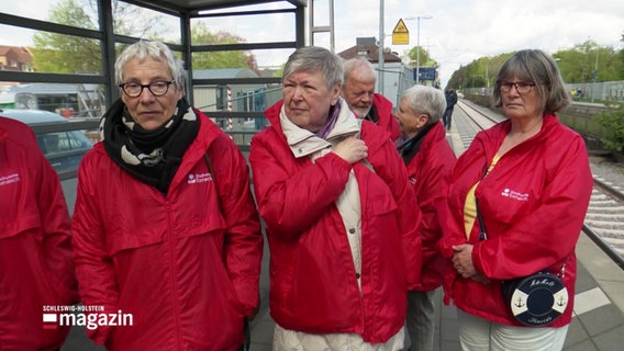 Damen in roten Jacken an auf einem Bahnsteig. © Screenshot 