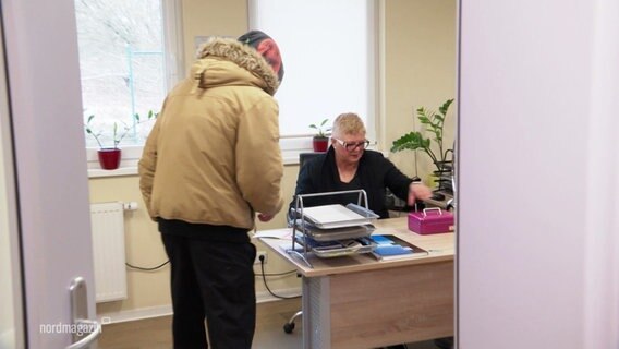 Ein Büro in dem eine Person am Schreibtisch sitzt und eine andere mit Kapuze gebückt davor steht. © Screenshot 