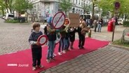 Schulkinder stehen mit Protestschildern gegen Autoverkehr und einem Hund auf einem roten Teppich, der über einen Zebrastreifen ausgebreitet liegt. © Screenshot 