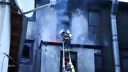Feuerwehrleute bei der Arbeit an einer verrußten Außenwand eines Gebäudes. © Screenshot 