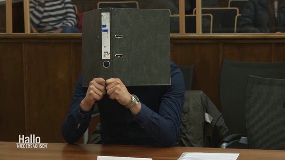 Ein Angeklagter hält sich eine Akte vor sein Gesicht. © Screenshot 