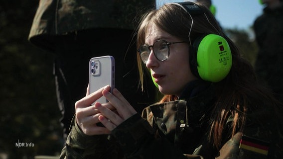 Ein Student in Tarnkleidung, mit Gehörschutz und Smartphone.  © Screenshot 
