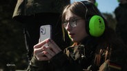 Eine Schülerin in Tarnkleidung, mit Gehörschutz und Smartphone. © Screenshot 